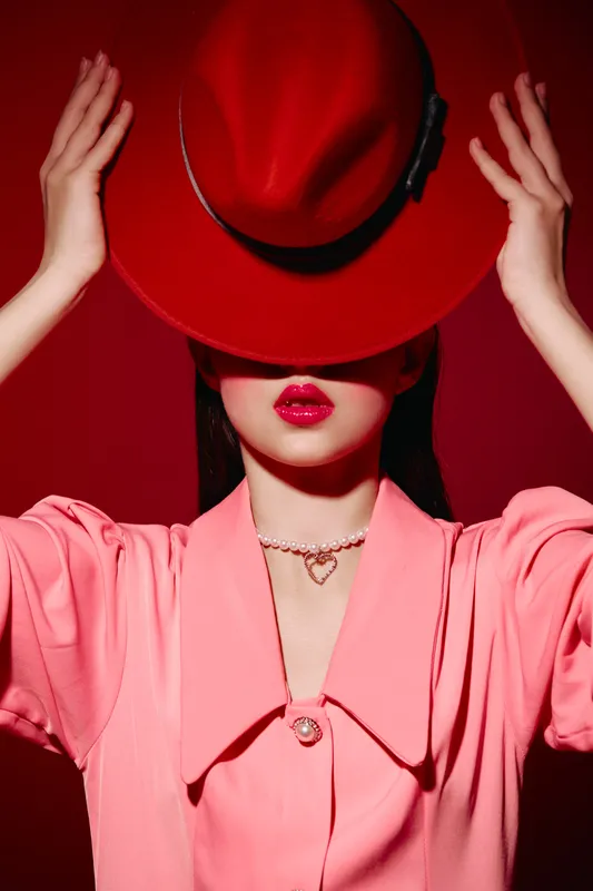 Stylish woman wearing a red hat and pink blazer poses against a red backdrop. Her face is mostly covered by the hat, highlighting red lips and a delicate necklace.