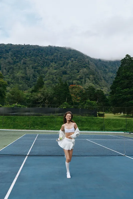Young woman in white tennis outfit joyfully running on a tennis court, surrounded by lush green trees and mountains, showcasing an active lifestyle and sporty vibe