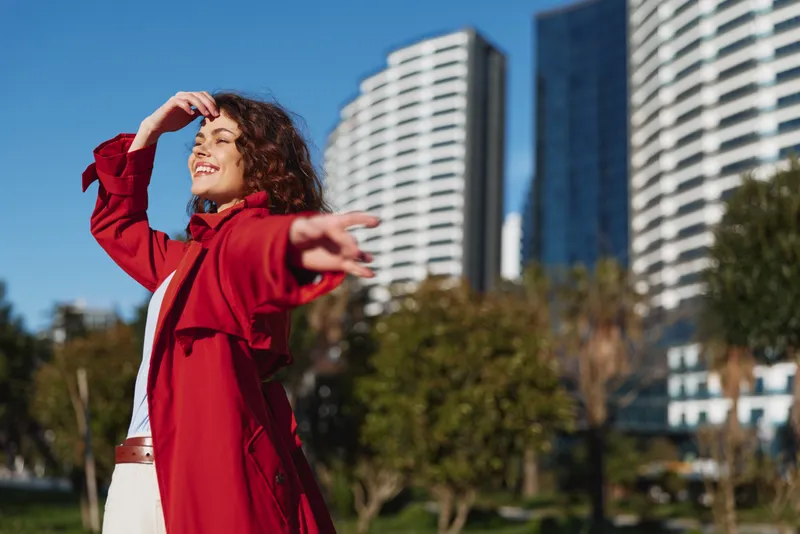Bright young woman outdoors in a red coat smiles with eyes closed, sunlit urban backdrop, carefree posture and a sense of freedom in a lively city moment with a gentle breeze