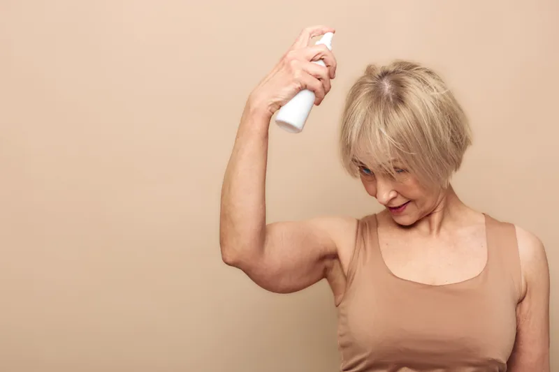 Mature woman with short blonde hair applies deodorant spray under her arm in beige tank top. Neutral background, personal hygiene and self-care concept for daily grooming routine.