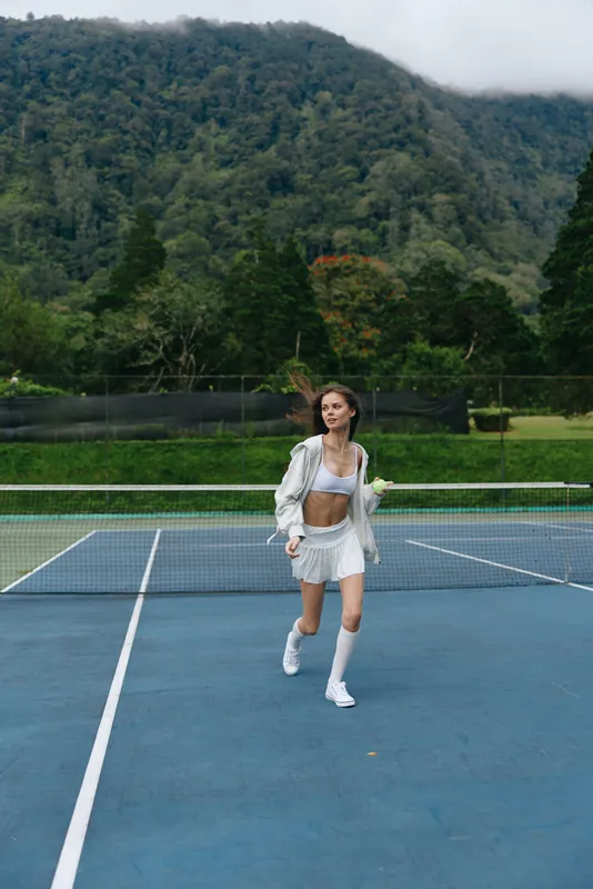 Young female tennis player running on a court, wearing a stylish white sports outfit, showing excitement and energy against a vibrant green mountainous background