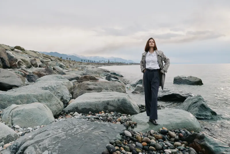 Woman stands on rocky coastline wearing casual jacket and trousers. Overcast sky and calm sea create moody atmosphere. Concept for nature, solitude, and outdoor lifestyle.