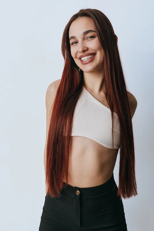 Young woman with long straight hair smiling and posing in casual outfit with white top and black pants against plain background. Happy female model, beauty and confidence.