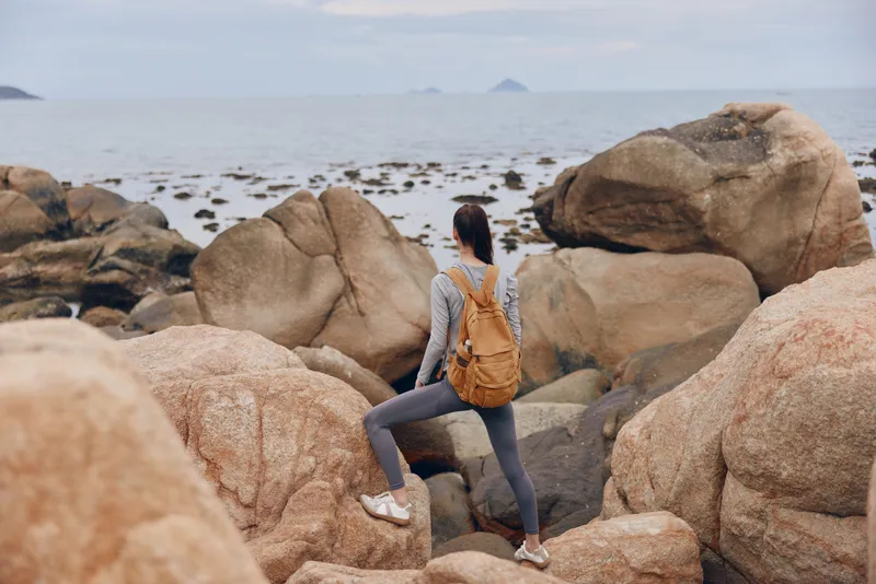 Young woman exploring rocky coastline with backpack, dressed in casual athletic wear, enjoying nature and adventure, perfect for outdoor activities