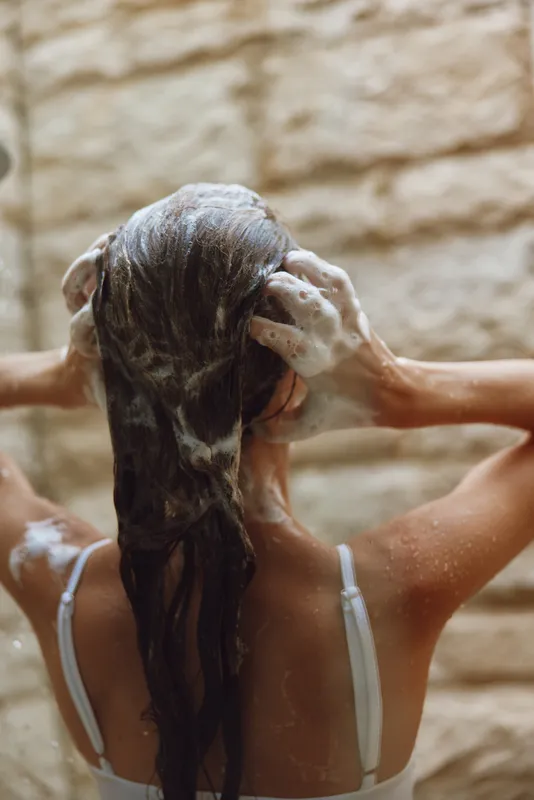 Woman washing long hair with shampoo, foam covering wet strands, in a casual white top against textured neutral background, calm and refreshing moment captured.