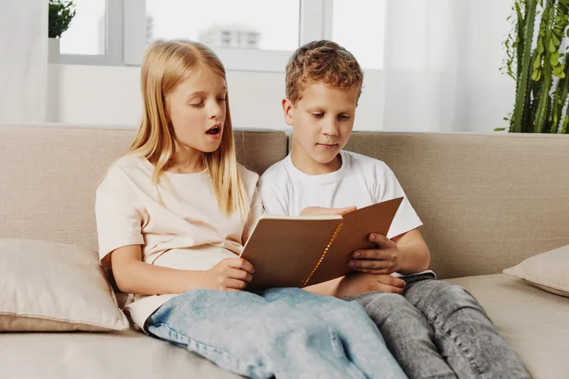 Two children sitting on a beige sofa reading a book together in a bright room. The girl and boy focus on the pages, sharing a quiet moment of learning and curiosity.