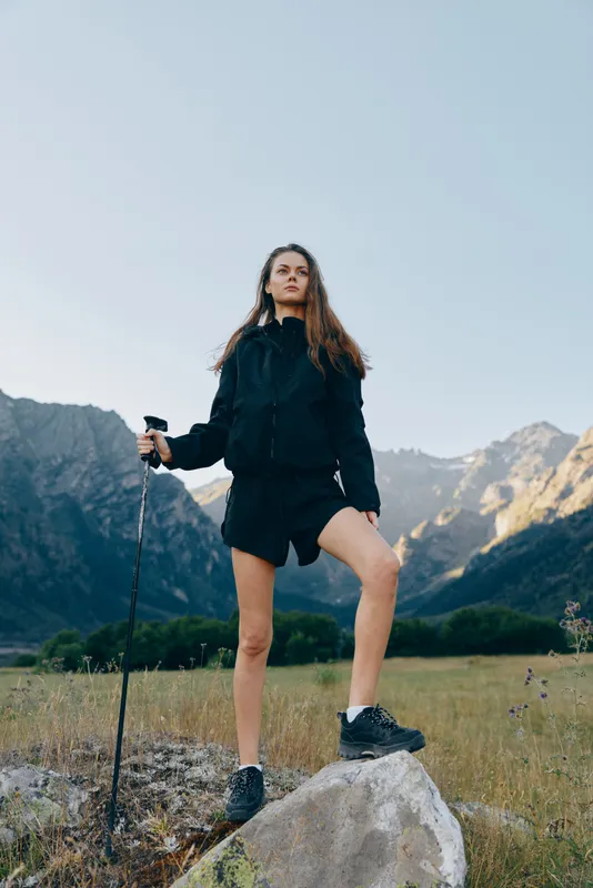 A focused hiker stands on a rock with a trekking pole in a mountain meadow. She gazes into the distance, exuding determination and a sense of adventure.
