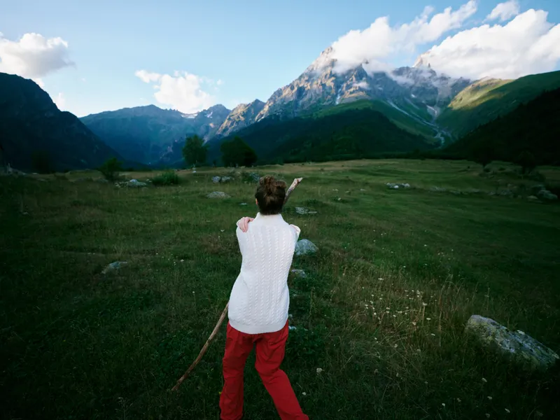 Back view of a hiker in a white top and red pants standing in a wide meadow with rocks, pointing toward distant mountains under a bright blue sky and clouds