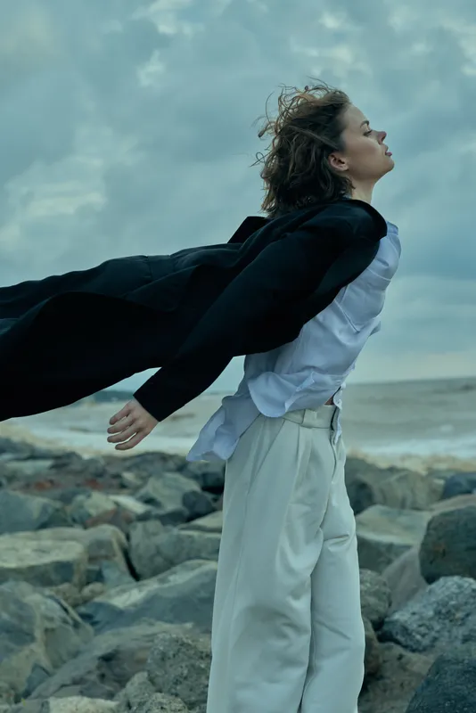 Wind sweeps around a figure on a rocky coast, cape unfurling, flowing white pants and dark jacket trailing behind, capturing bold movement and personal freedom against a windy shoreline