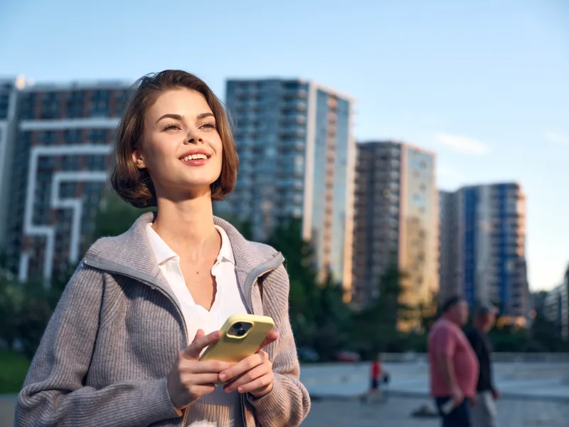 A confident professional woman stands outdoors in an urban setting, smiling while using a smartphone, with modern high rise buildings in the background.