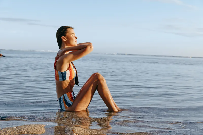 Relaxed woman in striped swimsuit sits on the beach shoreline with feet in shallow ocean water, arms behind her. Bright summer day beside calm sea, wellness concept.