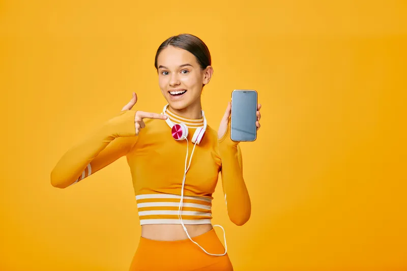 Fitness-themed portrait of a smiling woman in bright orange sportswear holding a smartphone and wearing headphones, ready for a workout in a vibrant studio backdrop.