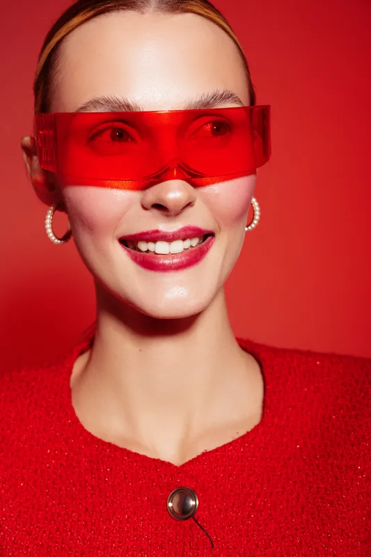 Smiling woman with red transparent glasses and red outfit poses on red background. Close-up portrait with glossy skin and hoop earrings. Concept of fashion, style, and modern accessories.