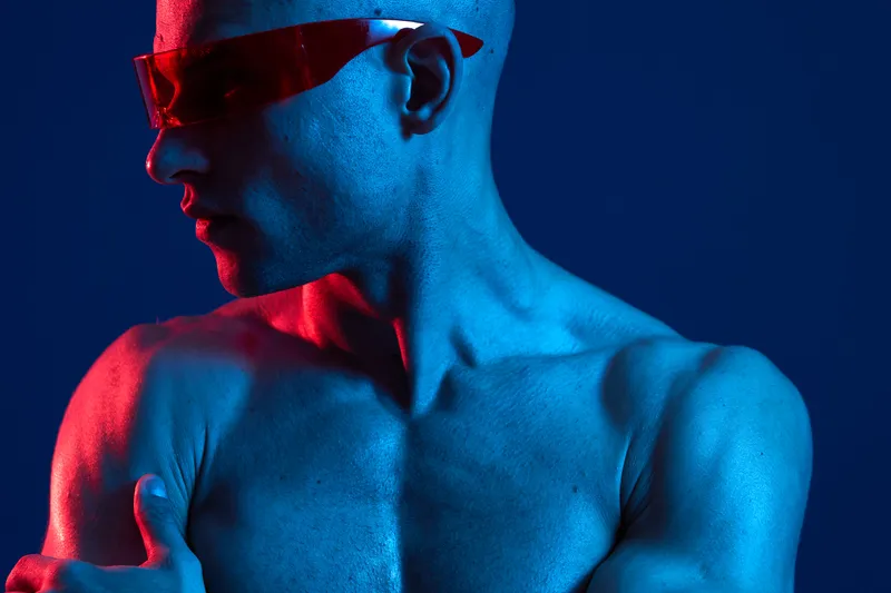 A stylish man showcases confidence with bold sunglasses, highlighting his muscular physique and vivid blue and red lighting, creating an energetic and dynamic atmosphere.