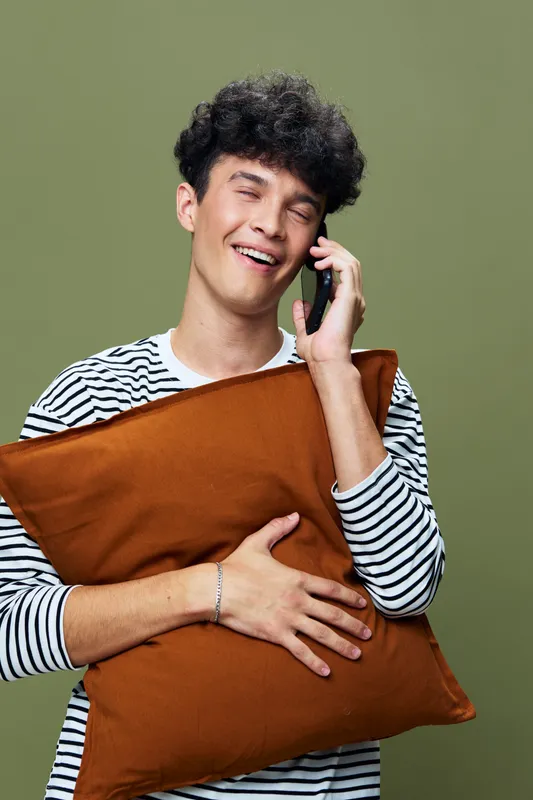 smiling man talking on phone holding brown pillow, casual striped shirt, curly hair, relaxed expression, olive green background, modern lifestyle concept