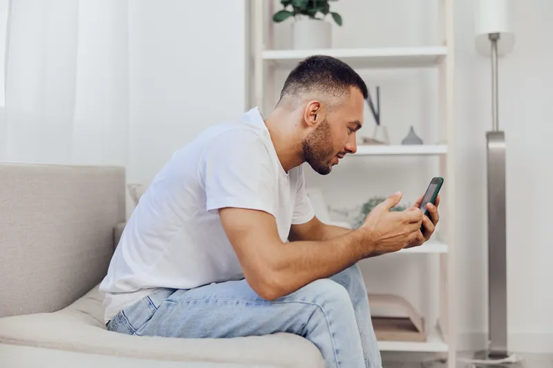 Focused man sitting on a couch, engaged with his smartphone, wearing casual clothing in a bright, modern living space, radiating concentration and intention.