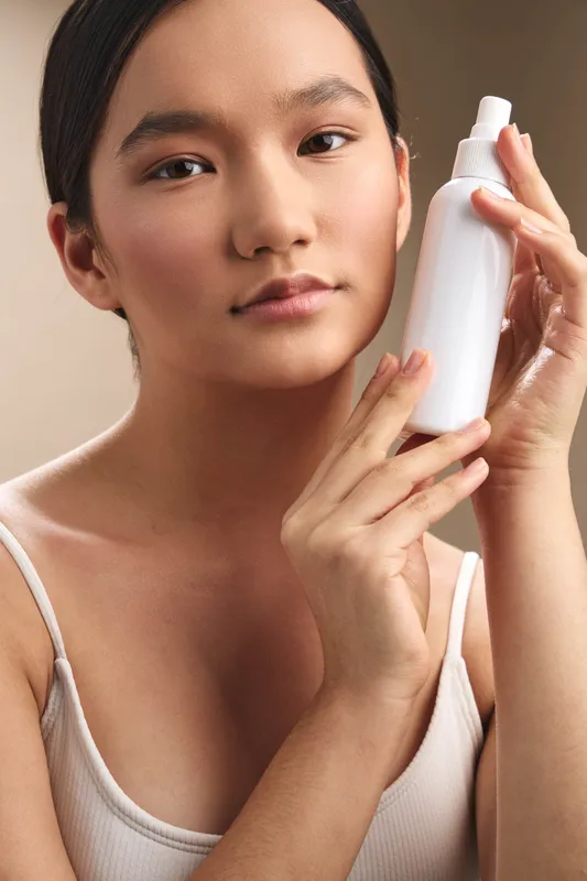 asian woman holds a cosmetic bottle in a soft studio setting, presenting skincare routine with calm confidence and a fresh minimal aesthetic.