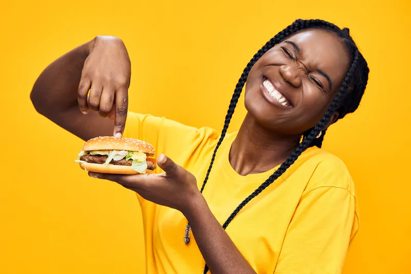 Happy young woman enjoying a delicious burger against a vibrant yellow background, showcasing the joy of healthy eating and fast food