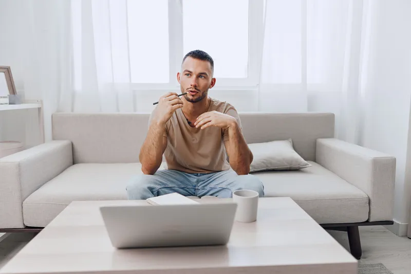 Thoughtful man sitting on a sofa, engaged in deep reflection while brainstorming ideas. Natural light filters through sheer curtains, creating a serene atmosphere.