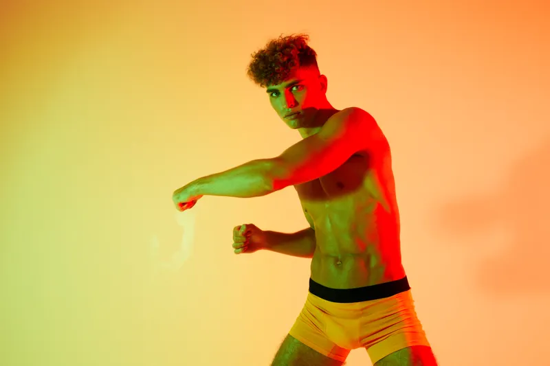 Young athletic man with curly hair posing shirtless in colorful lighting, showing muscular body and confident expression in studio with orange and green background.