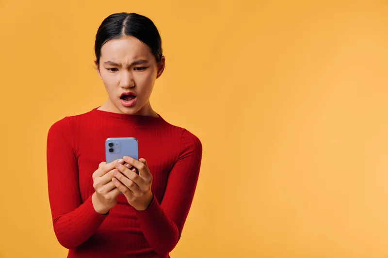 Young woman in red sweater looking at smartphone with shocked expression on yellow background, technology, communication, emotion, surprise, mobile device usage and reaction.