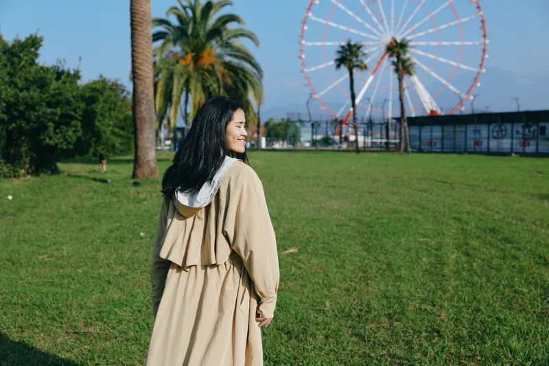 Park outdoor scene with a woman in a beige coat standing on grass, a ferris wheel in the distance, capturing leisure travel mood, serene vibe and exploration on a sunny afternoon amid greenery