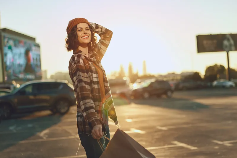 woman shopping smile golden hour candid urban authenticity lifestyle A young woman strolls through a sunlit parking lot with shopping bags, golden hour glow, candid lifestyle mood, mindful living and