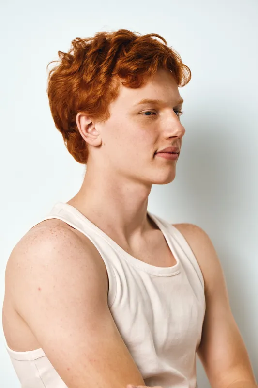 Young man with red curly hair wearing white tank top posing in studio profile view with crossed arms on white background. Casual confident male portrait with natural light.