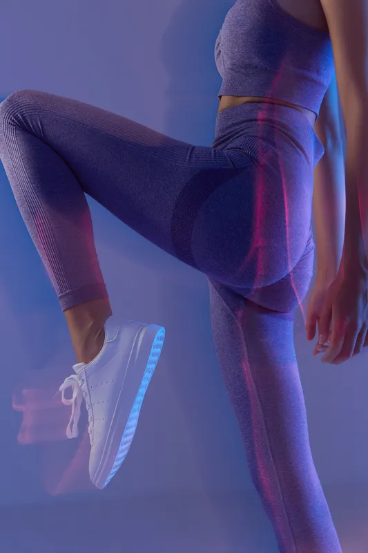 Fit woman sport fitness wearing purple leggings and white sneakers, exercising with leg raised indoors, dynamic pose and modern activewear style.