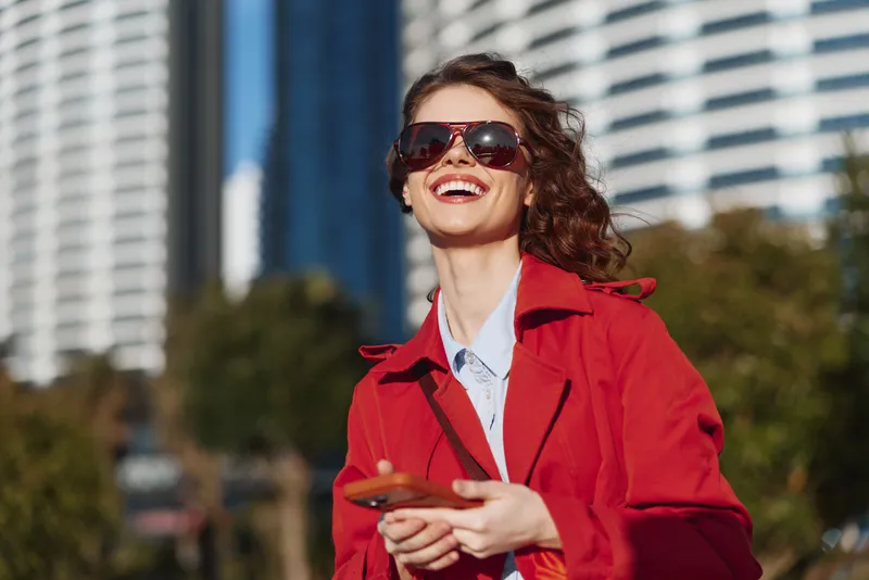 Smiling woman in a red coat wearing sunglasses, outdoors in an urban setting, bold fashion moment, bright daylight, confident mood, candid portrait.