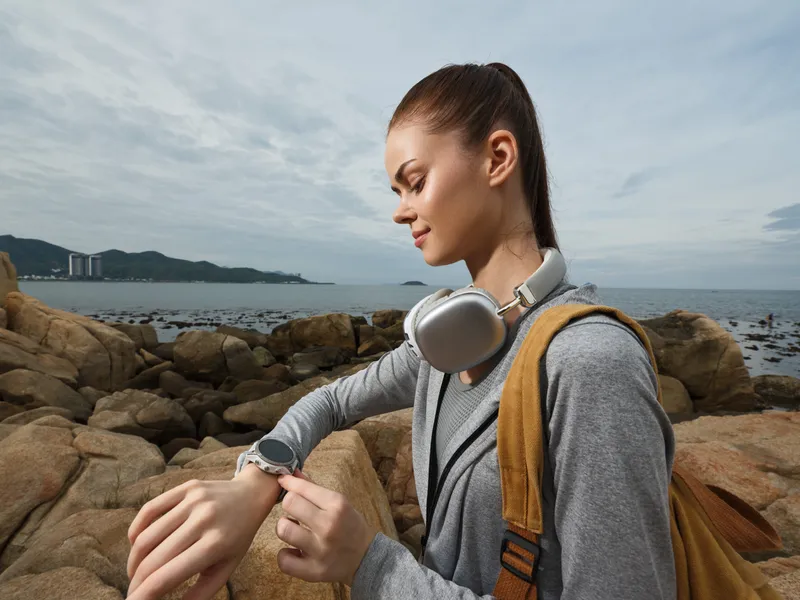 Young woman checking her smartwatch by the seaside, wearing a casual outfit and headphones, with a serene expression amidst a cloudy sky backdrop