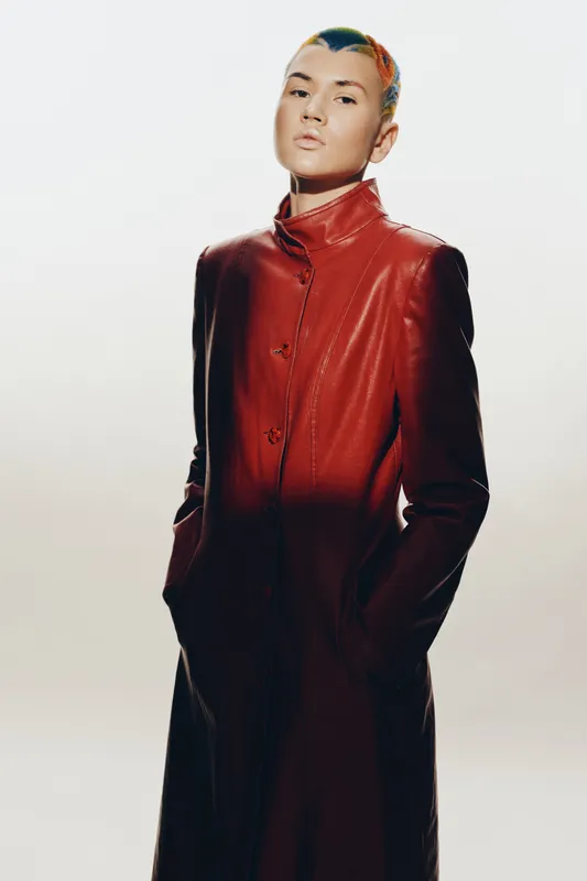 Woman with short colorful hair wearing a long red leather coat, posing confidently against a light neutral background with hands in pockets.