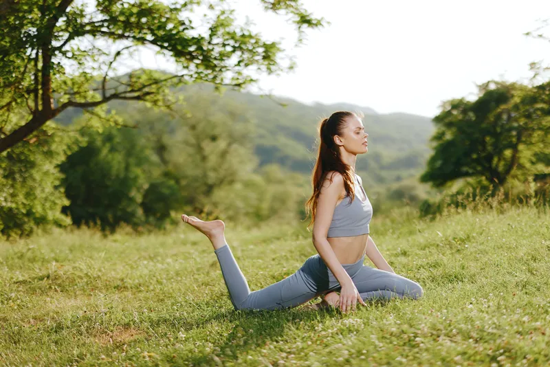 Serene young woman practicing yoga in a peaceful green landscape, wearing comfortable athletic clothing while enjoying the warm sun, emphasizing relaxation and mindfulness