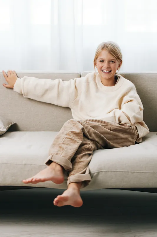 Authentic lifestyle shot of a cheerful child relaxing on a sofa in a cozy home, wearing a knit sweater and casual pants, centered on everyday comfort.