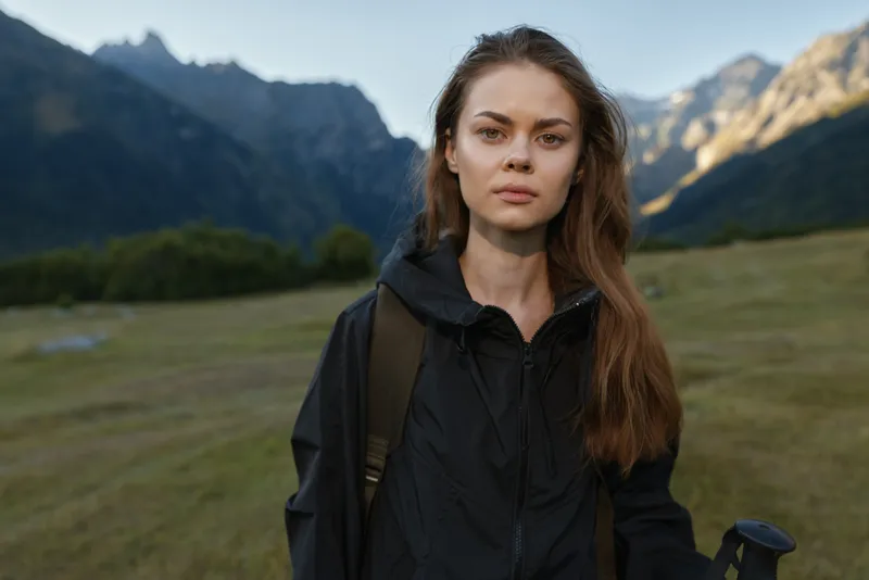 A confident traveler stands outdoors in a rugged mountain landscape, wearing a black jacket and backpack, looking directly at the camera with calm resolve and poised presence.