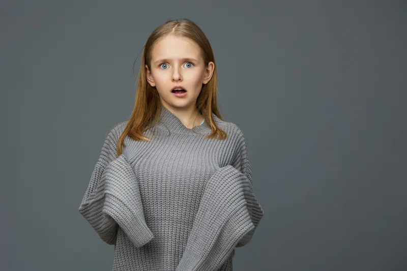 A surprised young girl in a cozy gray knitted sweater against a neutral background, capturing emotions of shock and curiosity, ideal for concepts of childhood and expression