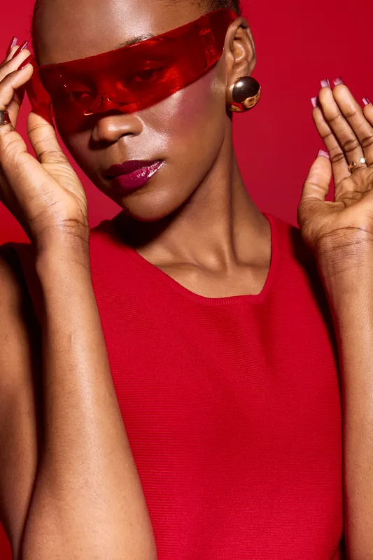 Close-up of elegant African woman with dark skin wearing red dress and red translucent glasses posing against red background. Concept of fashion, beauty, and style with vibrant colors.