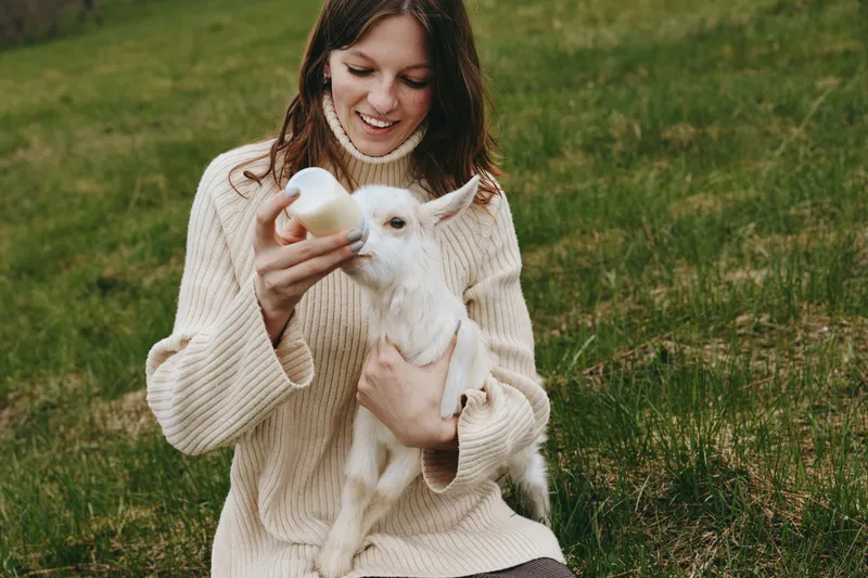 Goat kid shares a gentle feeding moment with a smiling woman outdoors, capturing a calm lifestyle and warm affection amid lush meadow greens, sunlit sky, and natural colors.