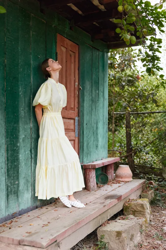 Woman in a pale yellow vintage dress poses outdoors by a rustic door, gazing upward with a serene garden backdrop, creating a timeless fashion moment full of charm and elegance
