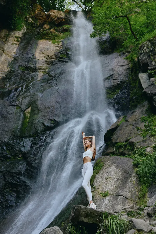young woman standing near waterfall in white activewear against rocky nature background, surrounded by lush green trees under natural daylight