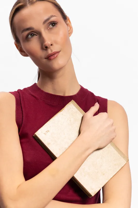 Young woman in red sleeveless top holds beige textured clutch purse, looking away thoughtfully. Minimalist style with white background. Concept for fashion, elegance, and contemplation.