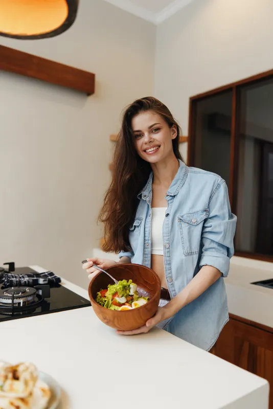 Счастливая молодая женщина смешивает свежий салат на стильной кухне, демонстрируя привычку к здоровому питанию и активный образ жизни