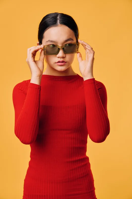 Woman in red long sleeve top adjusting sunglasses with both hands against yellow background looking forward with serious expression. Fashion, confidence and portrait of young adult female model.