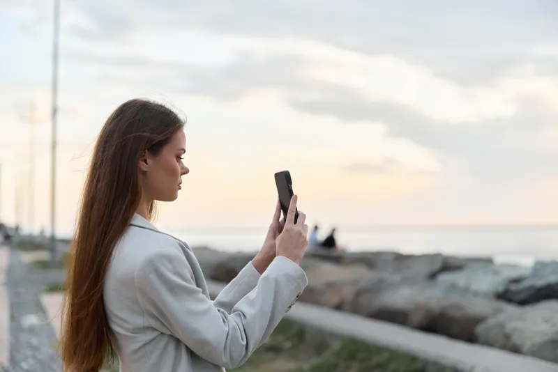 woman taking photo on smartphone outdoors during sunset with rocky coast background business concept
