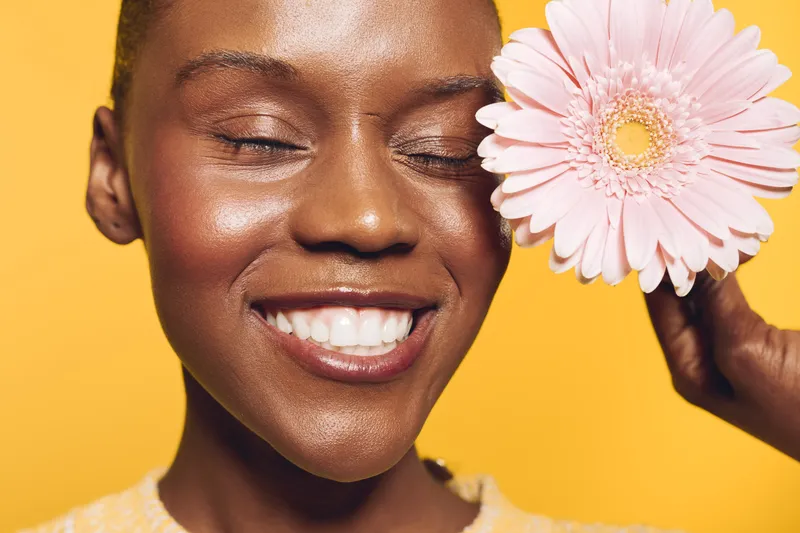 Close-up of smiling African woman with dark skin holding pink flower by face on yellow background. Concept of natural beauty, skincare, happiness, and wellness for black female model.