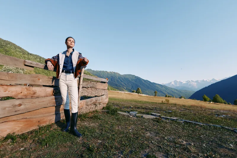 Woman leans on wooden fence in alpine meadow against distant mountains, countryside travel scene. Casual outfit with boots, relaxed portrait in wide open landscape under clear blue sky.