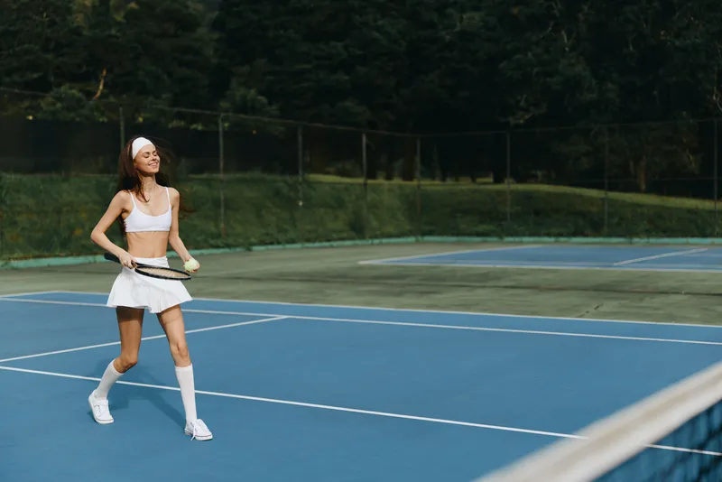 Young athletic woman playing tennis outdoors in a stylish white sport outfit, showcasing joy and energy on a blue court surrounded by nature