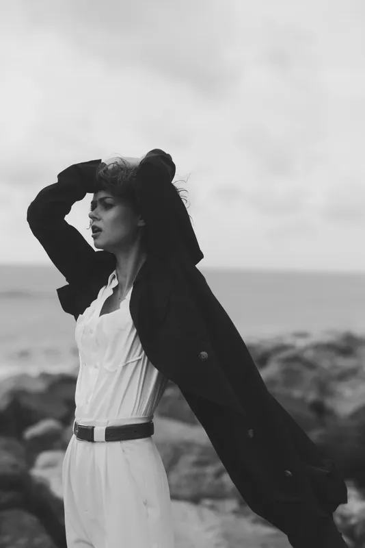 Woman on a windy coast wearing a white dress and dark coat, head tilted back, captured in monochrome for dramatic fashion and moody atmosphere.