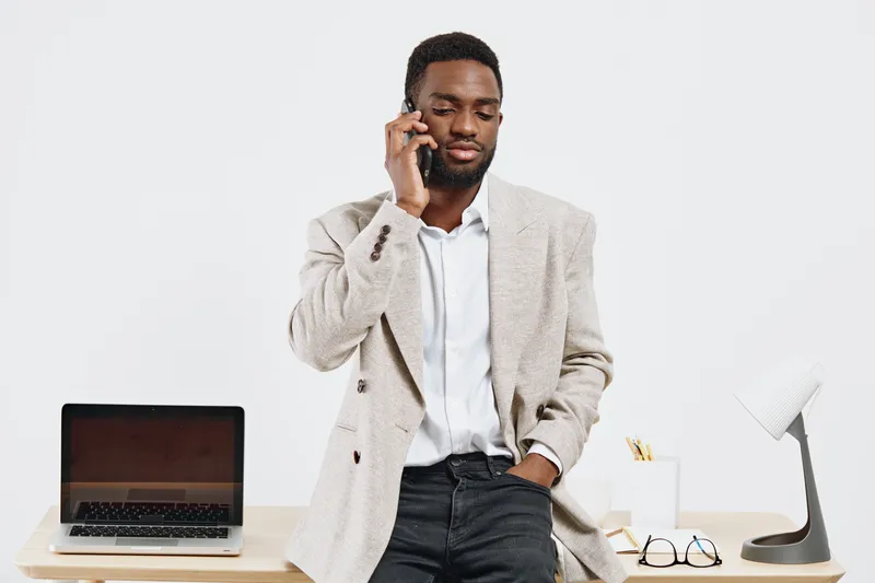 Professional young African American man in business casual attire talking on mobile phone while standing at office desk with laptop, glasses and writing tools, studio shot. Business communication conc