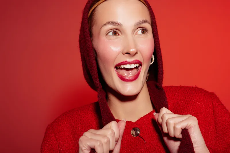 Happy woman in red hoodie smiles widely with bright lipstick on red background. Close-up portrait of cheerful female model with hoop earrings and natural skin glow. Concept of joy and fashion.